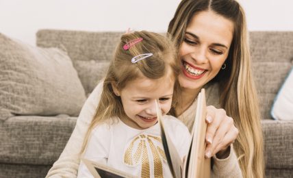 cheerful mother and daughter reading book near sofa
