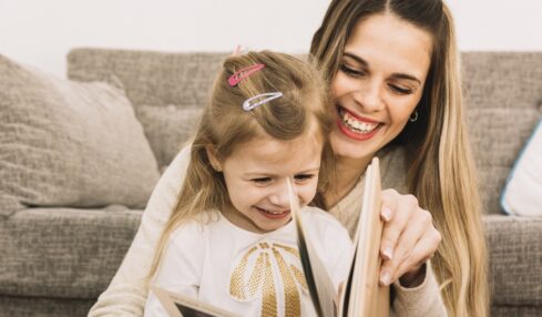 cheerful mother and daughter reading book near sofa
